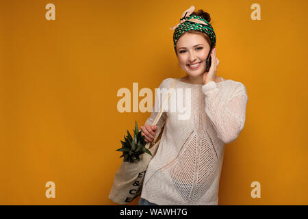 Jeune fille dans un foulard noué et pull léger avec un ananas dans un sac et le sourire au téléphone. Banque D'Images