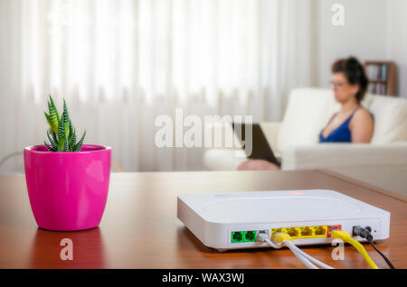 Modem routeur sur une table dans une salle de séjour avec une femme utilisant un ordinateur portable tout en étant assis sur un canapé en arrière-plan Banque D'Images