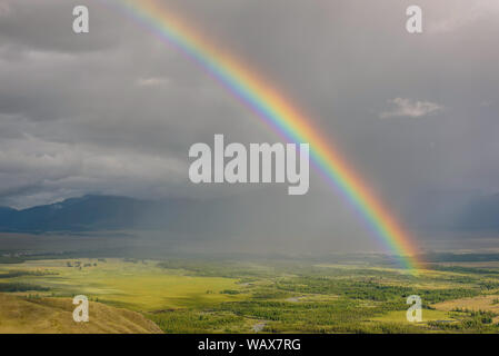 Arc-en-ciel de couleurs vives incroyable sur les montagnes, une vallée avec une rivière sinueuse et la forêt contre un ciel d'orage avec des nuages et de la pluie. L'Altaï, en Russie. Banque D'Images