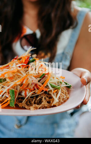 Woman holding dish of Thai street food cuisine - Som Tum salade de papaye Thaï épicé avec des frites croustillantes gâteau de poisson-chat Banque D'Images