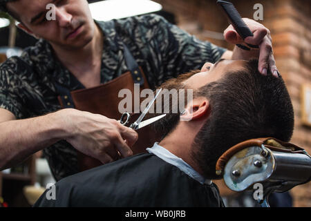 Close up portrait photo d'un salon de travail pour un beau gars à la coiffure. Il fait le style de sa barbe avec des ciseaux Banque D'Images