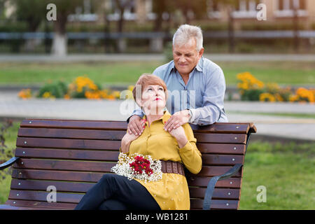 Une vieille femme est assise sur un banc et son mari se tient à côté d'elle et embrasse ses épaules. Banque D'Images