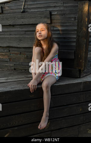 Pensive petit enfant aux cheveux longs reposant sur une terrasse en bois, à côté, tout en rêvant de quelque chose. Mignon bébé dans une robe blanche dans une Banque D'Images