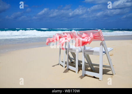 Chaises fixées sur une plage pour un mariage avec un ruban rose qui souffle dans la brise et les vagues lapant le fauteuil les jambes. Banque D'Images