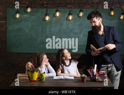 Enseignant et élèves filles en classe, tableau sur l'arrière-plan. Homme à barbe des écolières, enseigne la lecture de livre. Gai Écoute les enfants curieux Banque D'Images