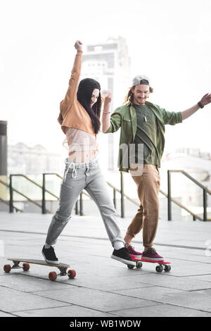 Couple heureux dans les vêtements de se tenir la main, à cheval sur la planche à roulettes dans la ville Banque D'Images