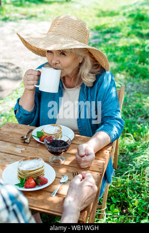 Senior woman in straw hat à boire le thé près de sweet et délicieuses crêpes Banque D'Images