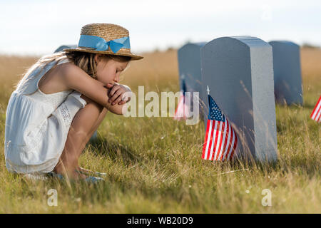 Enfant triste en chapeau de paille assis près de pierres tombales avec american flags Banque D'Images