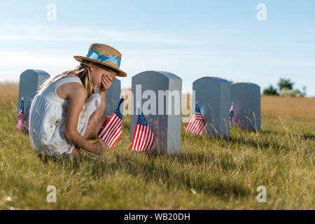 Kid en chapeau de paille couvrant visage tout en s'asseyant à côté de pierres tombales avec american flags Banque D'Images