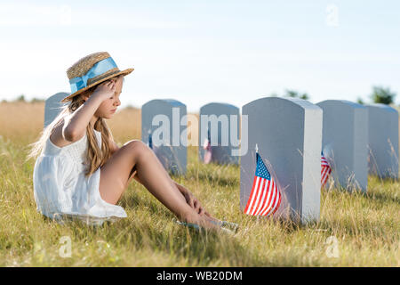 Kid en chapeau de paille donnant alors que saluer assis près de pierres tombales avec american flags Banque D'Images