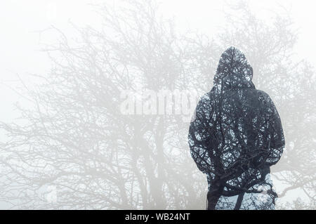 Une double exposition, à à l'arrière d'une silhouette encapuchonnée plus de couches par branches d'arbres en hiver. Banque D'Images