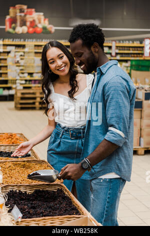 Happy african american man holding metal écope avec raisins secs près de asian woman gesturing in store Banque D'Images