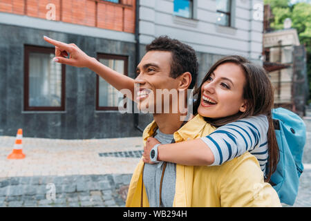 Happy girl hugging cheerful mixed race man et en pointant avec le doigt près du bâtiment Banque D'Images