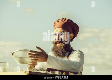 Chef cuisinier homme hipster avec beau visage expressif barbu riant dans hat et blanc et rouge farine tamis avec boulonnage uniforme sur fond de ciel bleu Banque D'Images