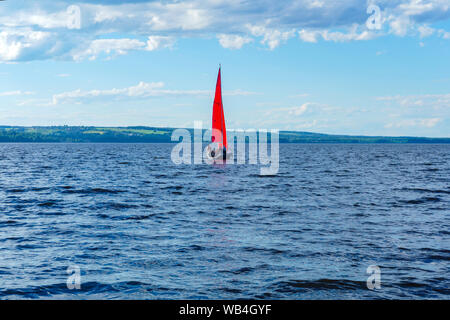 Petit voilier avec voiles rouge au milieu d'une vaste étendue d'eau avec une rive boisée à l'horizon Banque D'Images