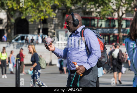 Bald Middle aged black man walking in Summer portant des écouteurs et grande à l'aide d'un smartphone dans le centre de Londres, Angleterre, Royaume-Uni. Banque D'Images
