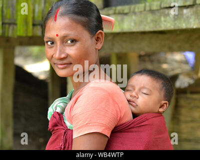 Les Indiens mariés Mishing femme tribal (sindoor rouge et bindi sur le front) de l'Île Majuli porte son sleeping baby boy dans un porte-bébé traditionnel. Banque D'Images