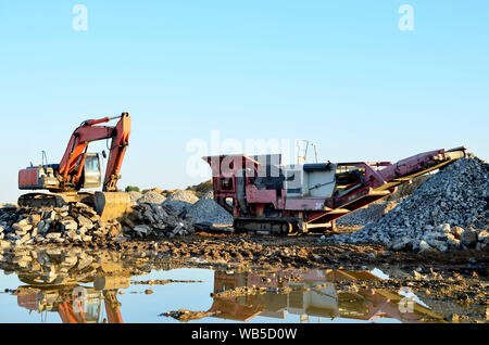 Excavatrice lourde travaillant dans une carrière sur un fond de coucher de soleil et ciel bleu. Concasseur à mâchoires mobile par le site de construction. Vieux béton concassage a été Banque D'Images