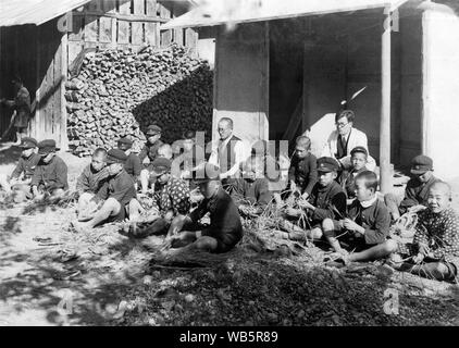 [ 1930 - Japon ] Les élèves de l'école primaire japonaise - les enfants de l'école élémentaire des hommes japonais en uniforme assis sur le sol à côté d'un bâtiment de ferme font des sandales de paille (waraji). c1920s ou 1930. 20e siècle Tirage argentique d'époque. Banque D'Images