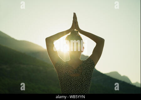 Vue de derrière d'une jeune femme faisant asana yoga avec le soleil du matin brille à travers un écart entre son bras et la tête. Banque D'Images