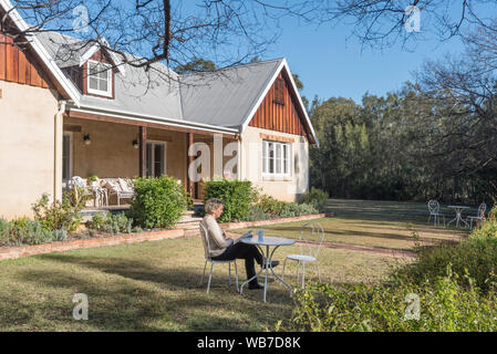 Une femme blanche d'âge moyen se situe à un cadre extérieur blanc en hiver matin soleil dans un jardin devant une maison avec une tasse de thé en lisant un livre Banque D'Images