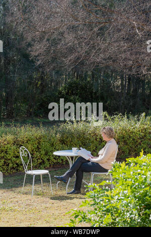 Une femme blanche d'âge moyen est assis à une table et chaises extérieur blanc en hiver matin soleil dans un jardin avec une tasse de thé en lisant un livre Banque D'Images