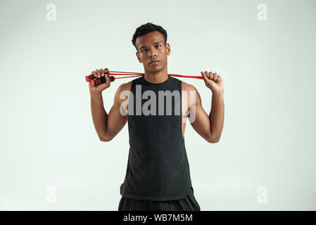 Prêt pour l'entraînement. Strong et sportive homme jeune tenant une corde à sauter dans son dos et looking at camera while standing against fond gris. Les équipements de sport. Mode de vie actif Banque D'Images