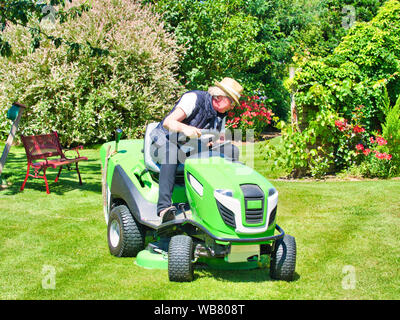 Woman hat conduisant un tracteur tondeuse de jardin avec fleurs. En vert et blanc sur la faucheuse, virage dans le champ entre fleurs colorées. C Banque D'Images