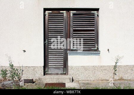 Maison de la famille les portes d'entrée avec fenêtre en bois délabrées fermé avec stores décolorées monté sur façade gris clair entouré de béton Banque D'Images