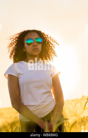 Belle mixed race girl biracial African American female adolescent jeune femme portant des lunettes de soleil au coucher et au lever du soleil dans un champ de cultures Banque D'Images