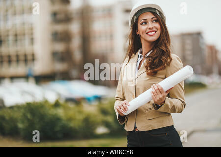 Belle jeune femme ingénieur avec casque blanc et les bleus dans ses mains en face de l'office de district. Banque D'Images