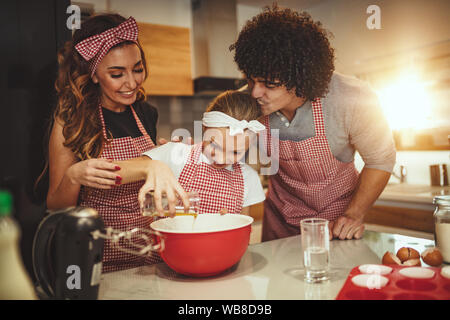 Heureux parents et leur fille sont en train de préparer des cookies ensemble dans la cuisine. Petite fille à ses parents aide à verser l'huile dans le plat avec la pâte. Banque D'Images