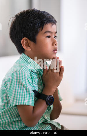 Mixed Race jeune garçon tient ses mains en prière et assis sur la chaise Banque D'Images