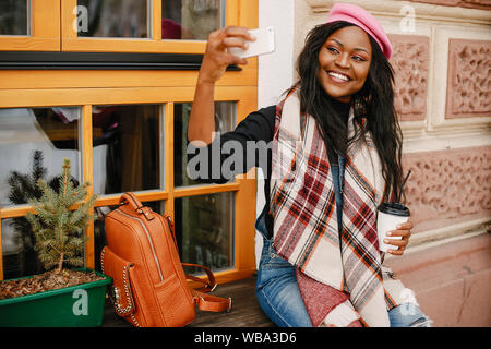 Cute black woman s'amuser dans une ville. Belle fille noire dans un béret rose. Dame élégante de boire un café. Mort a utiliser le téléphone Banque D'Images