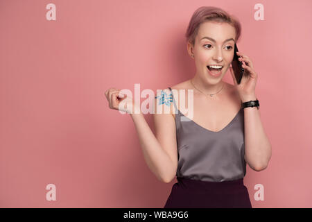 Une fille avec de courts cheveux roses et un tatouage sur son épaule dans un haut gris parle avec enthousiasme sur le téléphone sur un fond rose. Banque D'Images