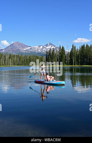 Deux jeunes femmes debout sur paddle board sur Scott Lake, Oregon avec centre et nord volcans en arrière-plan. Soeurs Banque D'Images