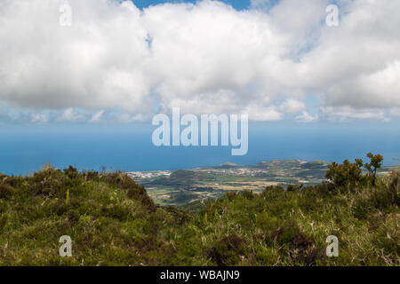 Les petites collines couvertes d'un champs et prés verts frais, relevant le niveau de l'océan Atlantique à l'arrière-plan. Ciel bleu avec des nuages blancs intenses Banque D'Images