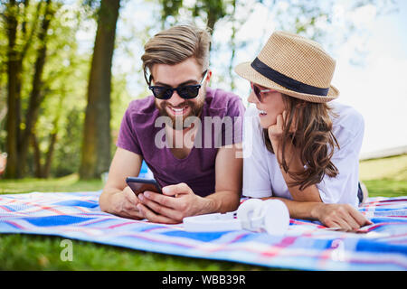 Jeune couple aimant tout en ayant l'aide de Phone lying on blanket et pique-nique en été Banque D'Images