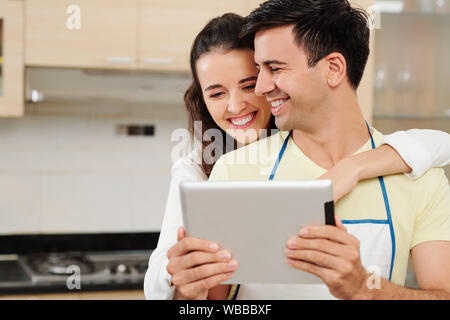 Pretty smiling young woman hugging son petit ami par derrière et pointant sur écran de tablette numérique dans les mains quand qui sont debout dans la cuisine Banque D'Images