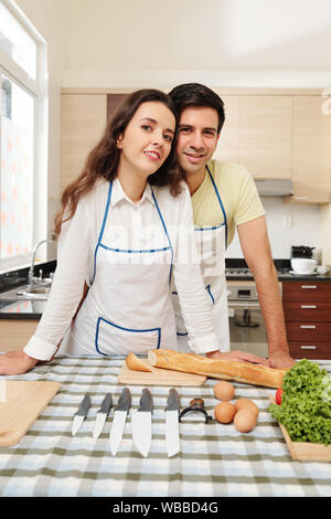 Cheerful young couple en tablier blanc debout à leur table de cuisine and smiling at camera Banque D'Images