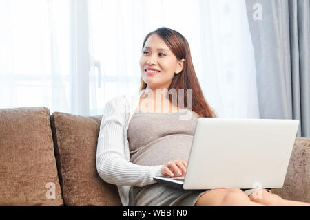 Smiling young pregnant Asian woman working on laptop lorsque sitting on sofa at home Banque D'Images
