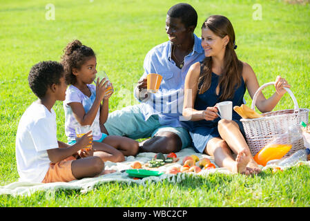 Happy mixed race couple avec enfants préadolescentes gaiement passant le temps à pique-niquer sur la pelouse verte Banque D'Images