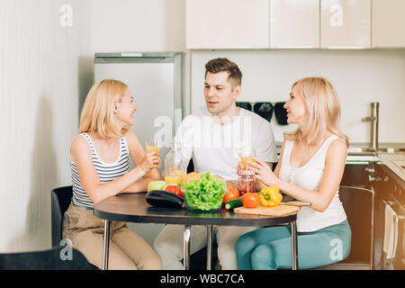Amis aller à préparer des aliments sains à partir de légumes. Deux jolies femmes heureux et bel homme assis dans la cuisine, boire du jus et de parler. Lifesty Banque D'Images