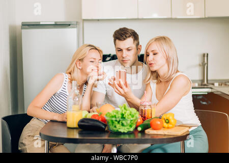 Portrait of cheerful young friends looking at smartphone tout en étant assis à table dans la cuisine. Deux belles femmes blondes et l'homme à l'aide de téléphone mobile, watchi Banque D'Images