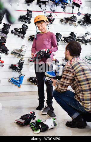 Jeune homme souriant satisfait vendeur aider boy en essayant sur patins à roulettes dans magasin de sport Banque D'Images