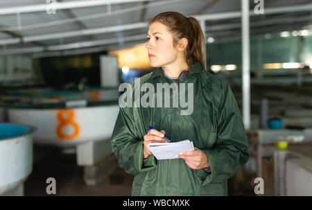 Portrait de femme de l'inspection des réservoirs pour les alevins de truite d'élevage dans les exploitations agricoles, en prenant des notes dans l'ordinateur portable Banque D'Images