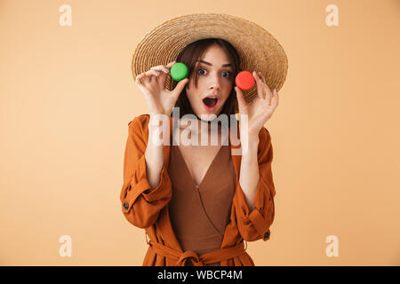 Belle jeune femme portant chapeau de paille et de l'été tenue isolés sur fond beige, tenant deux macarons Banque D'Images