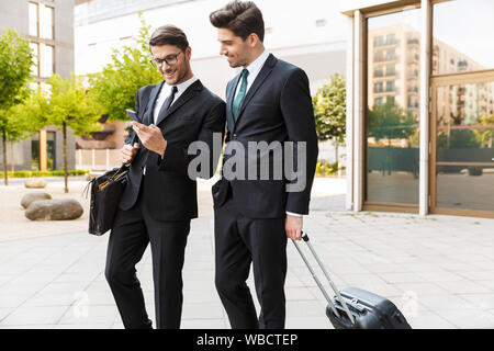 Deux beaux jeunes hommes d'souriant portant des costumes à la marche à l'extérieur les rues de la ville, transportant valise, using mobile phone Banque D'Images