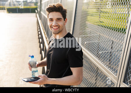 Portrait of smiling man in sportswear holding cellulaire et une bouteille d'eau en se tenant sur le terrain de sport au cours d'exercices du matin à l'extérieur Banque D'Images