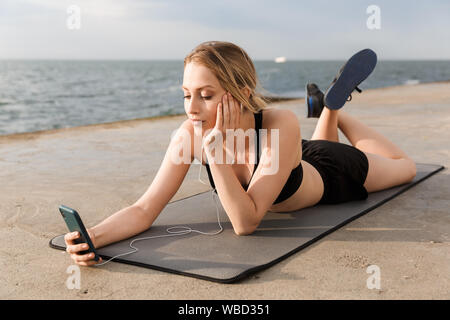 Image de jeune femme portant ensemble en écoutant de la musique sur téléphone portable tout en étant allongé sur le tapis près de mer à matin Banque D'Images